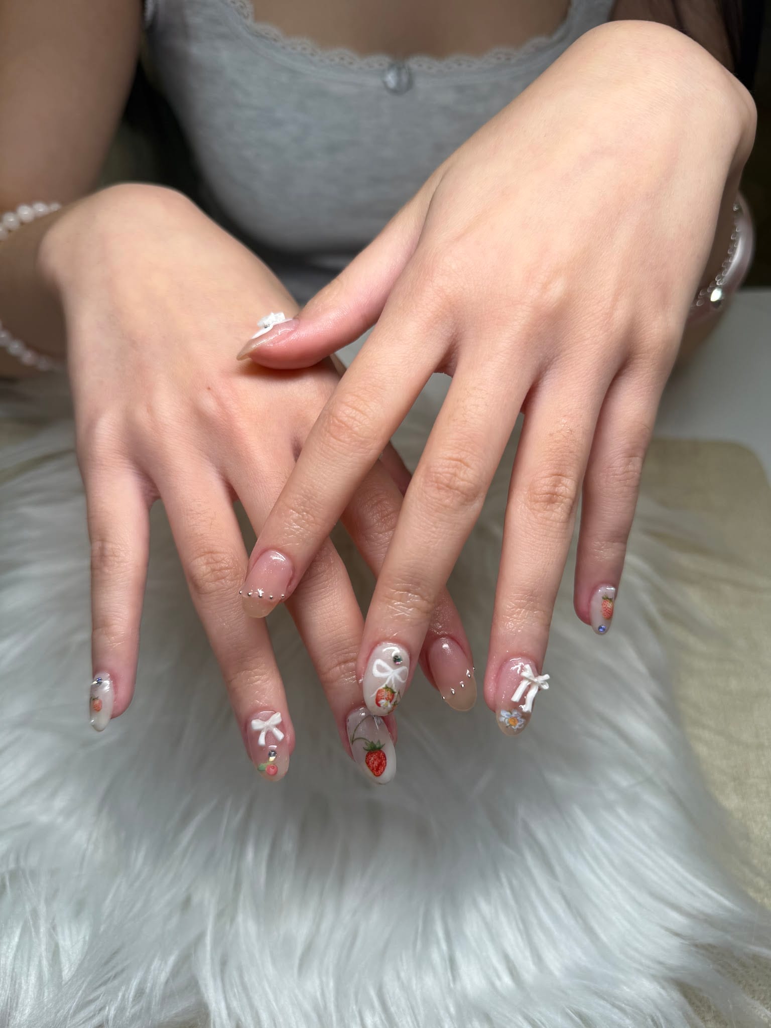 Hands displaying acrylic nails with strawberry art, white bows, and rhinestones