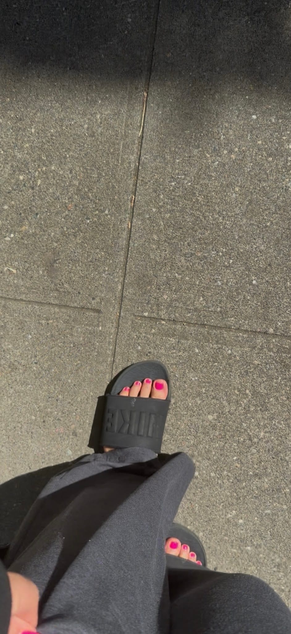 Feet with bright pink pedicure in black Nike slides on concrete